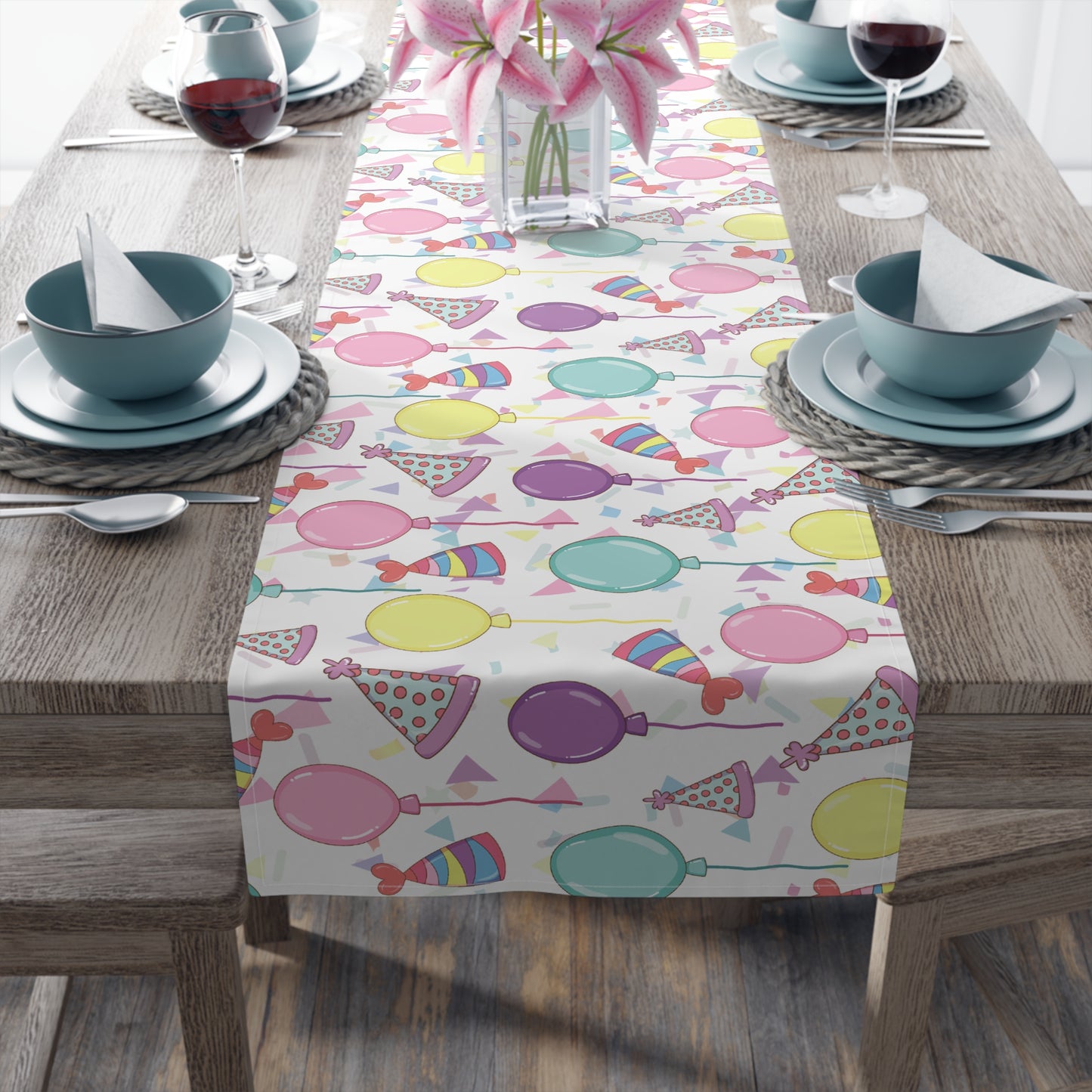 Table runner with birthday hats, confetti and purple, yellow, blue and pink balloons design on a white background on a dark wood table with blue plates and bowls