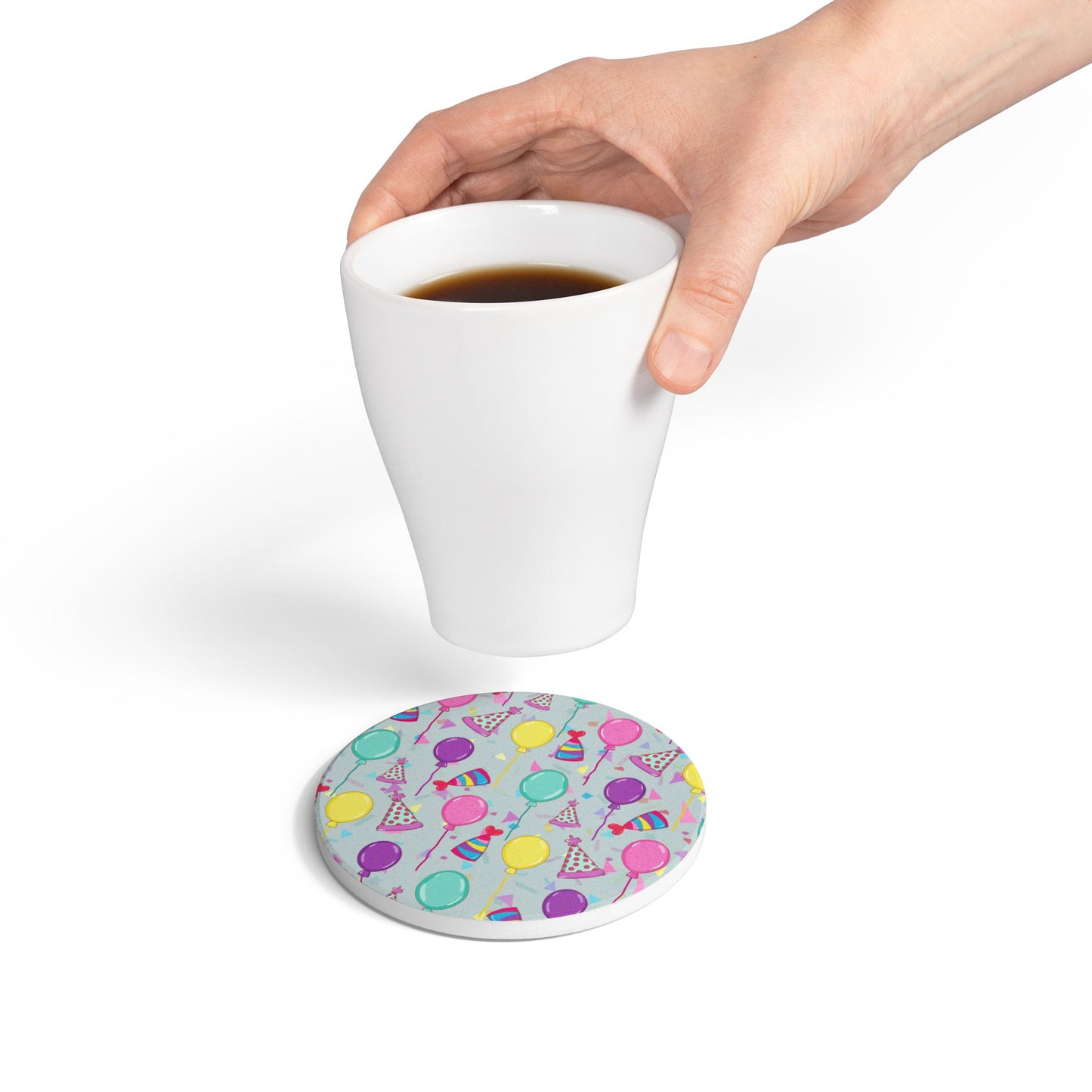 Happy Birthday round ceramic tile coaster with with birthday hats, confetti and balloons pattern with a blue-green light cyan background on white background with person placing coffee cup on coaster