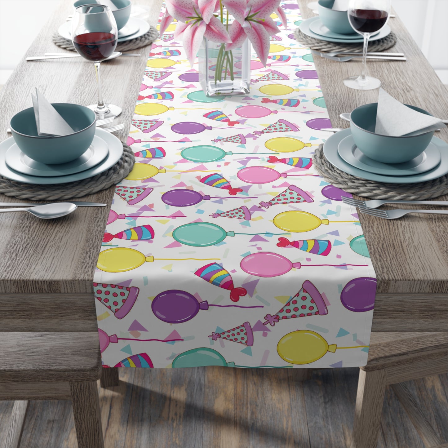 Table runner with birthday hats, confetti and purple, yellow, blue and pink balloons on a white background on a dark wood table with blue plates and bowls