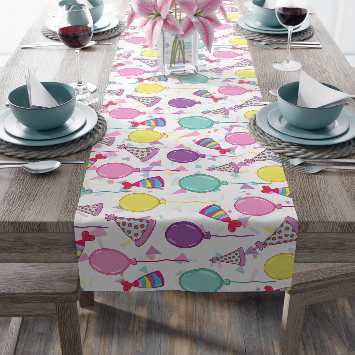 Table runner with birthday hats, confetti and purple, yellow, blue and pink balloons design on a white background on a dark wood table with blue plates and bowls