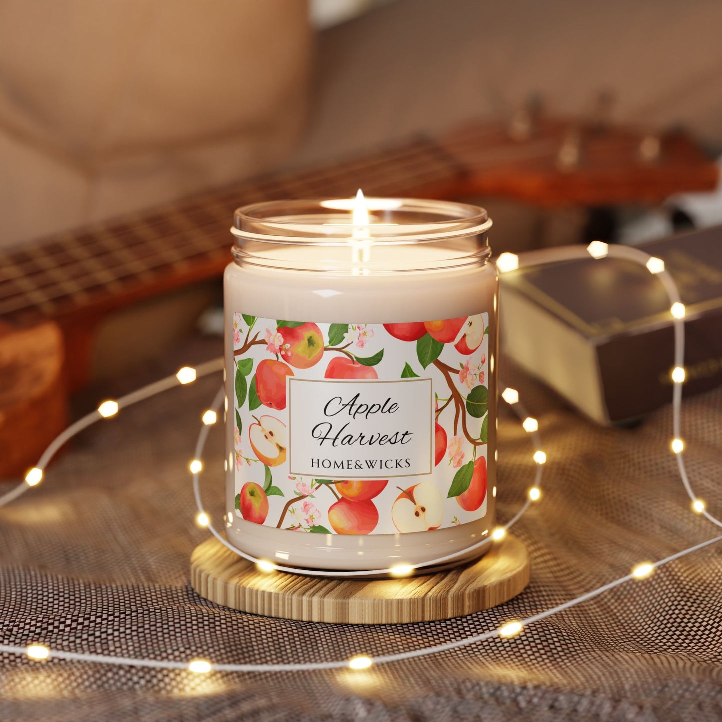 Lit candle in a jar with 'Apple Harvest' label on a wooden coaster, surrounded by string lights and a guitar in the background.