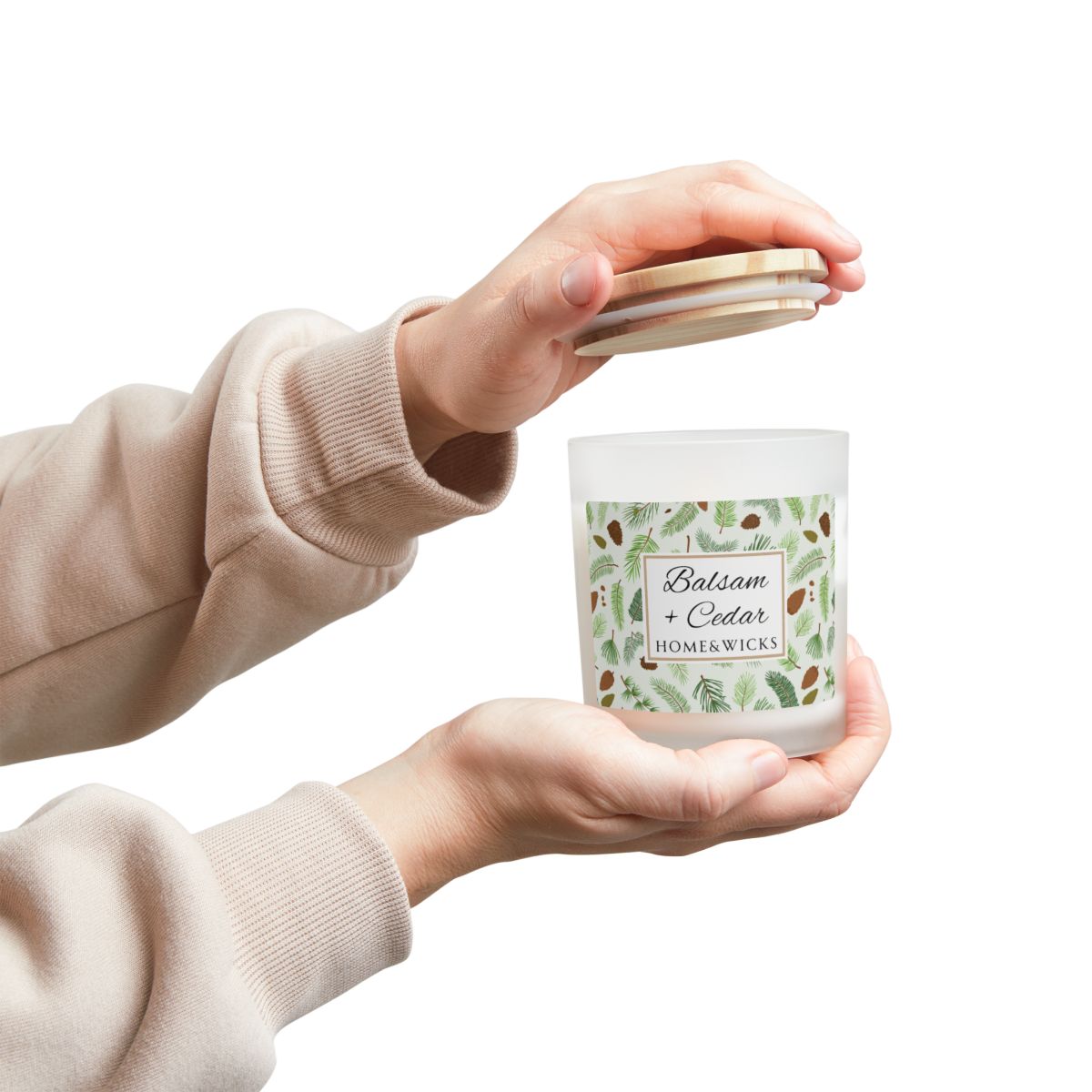 Wood wick candle with 'Balsam + Cedar' label with cones and cedar leaves on a white background by Home & Wicks with Person holding a candle labeled 'Balsam + Cedar' by Homer Wicks on a white background
