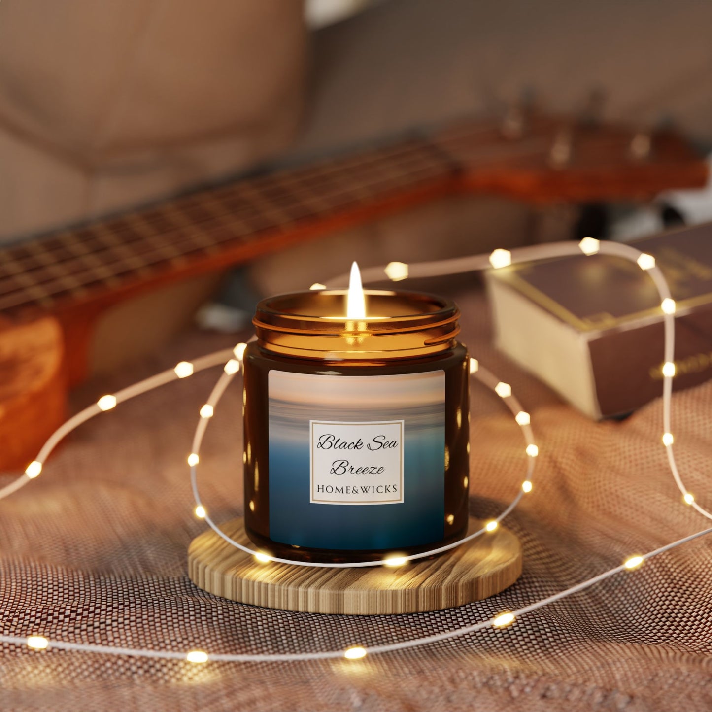 Candle labeled 'Black Sea Breeze' by Home & Wicks on a wooden coaster with string lights and a guitar in the background.
