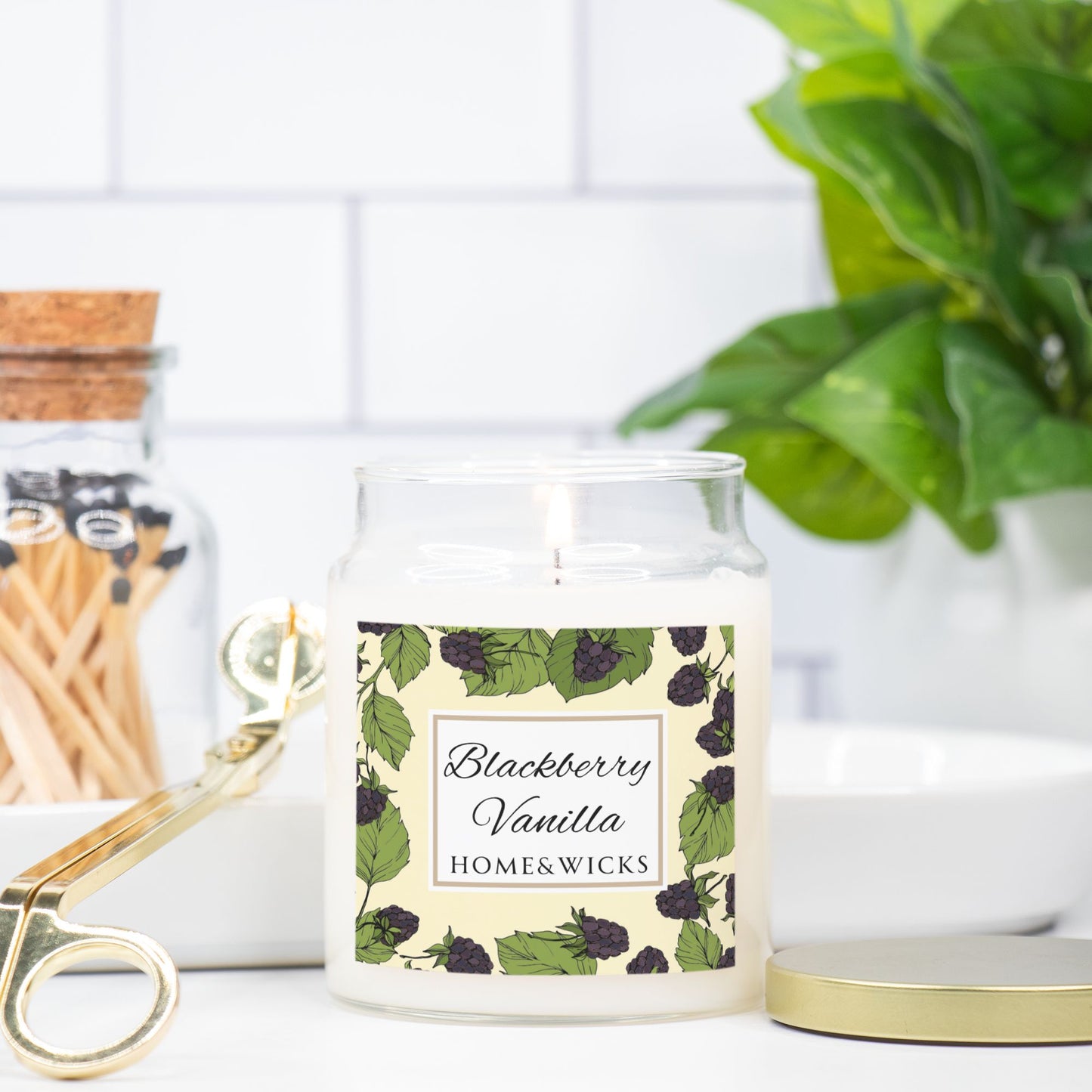 Candle jar labeled 'Blackberry Vanilla' by HOME & WICKS on a white surface with a plant and matches in the background.