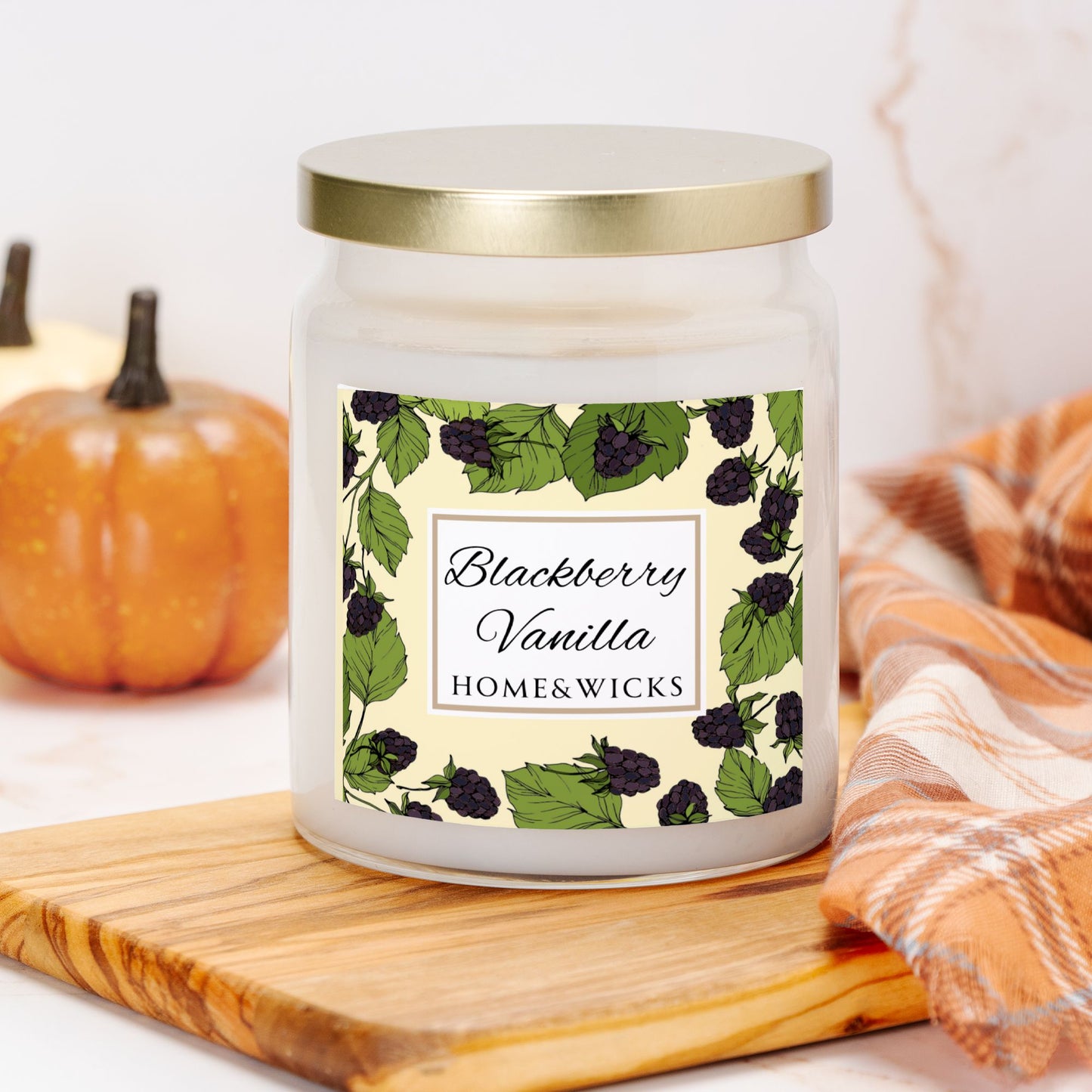 Candle with a blackberry vanilla label on a wooden board with pumpkins and a blanket in the background