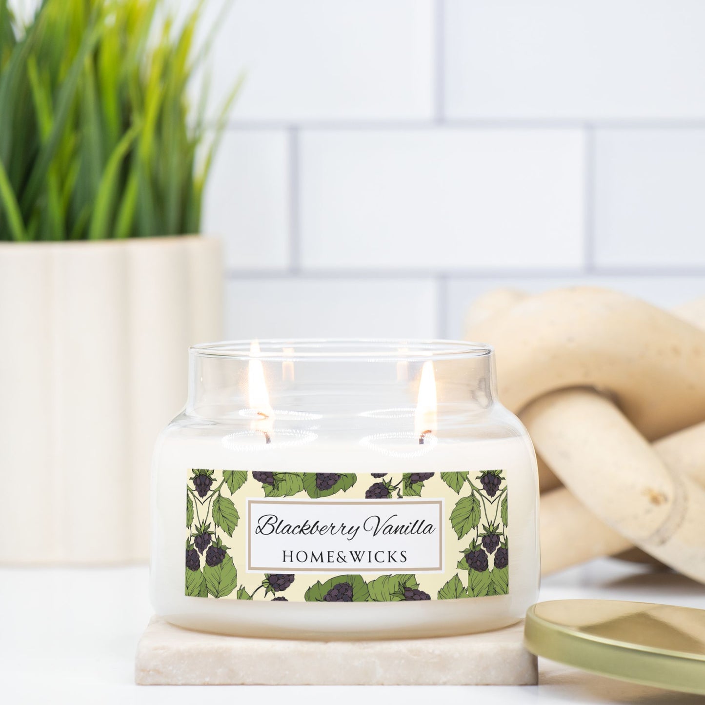 Candle with a 'Blackberry Vanilla' label on a marble surface, with a plant and wooden logs in the background.