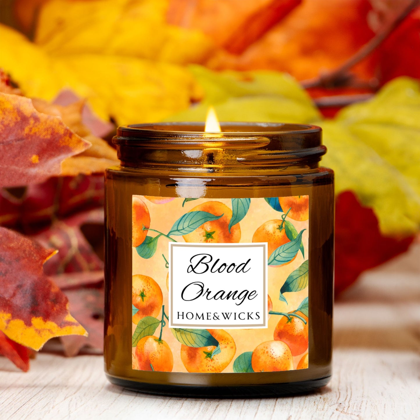 Blood Orange candle in an amber glass jar with gold lid with 'Blood Orange' label featuring oranges illustration on a white background by Home & Wicks with autumn leaves in the background