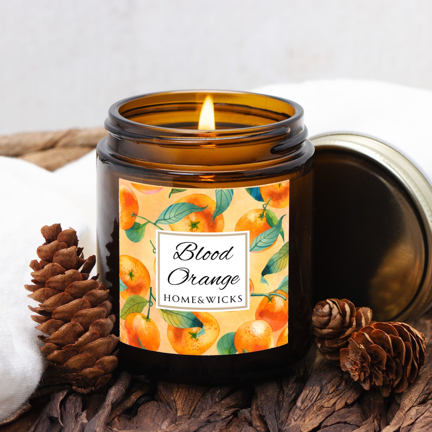 Blood Orange candle in an amber glass jar with gold lid with 'Blood Orange' label featuring oranges illustration on a white background by Home & Wicks with decorative pine cone elements on a neutral background