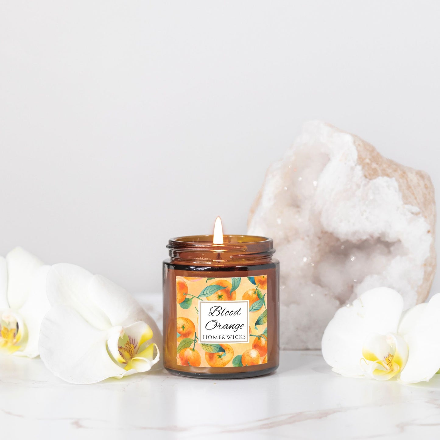 Blood Orange candle in an amber glass jar with gold lid with 'Blood Orange' label featuring oranges illustration on a white background by Home & Wicks on a white surface with decorative orchid elements