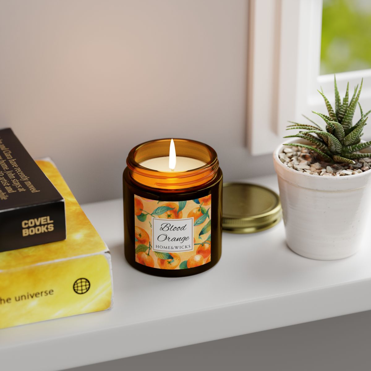 Blood Orange candle in an amber glass jar with gold lid with 'Blood Orange' label featuring oranges illustration on a white background by Home & Wicks books, and a plant on a surface.
