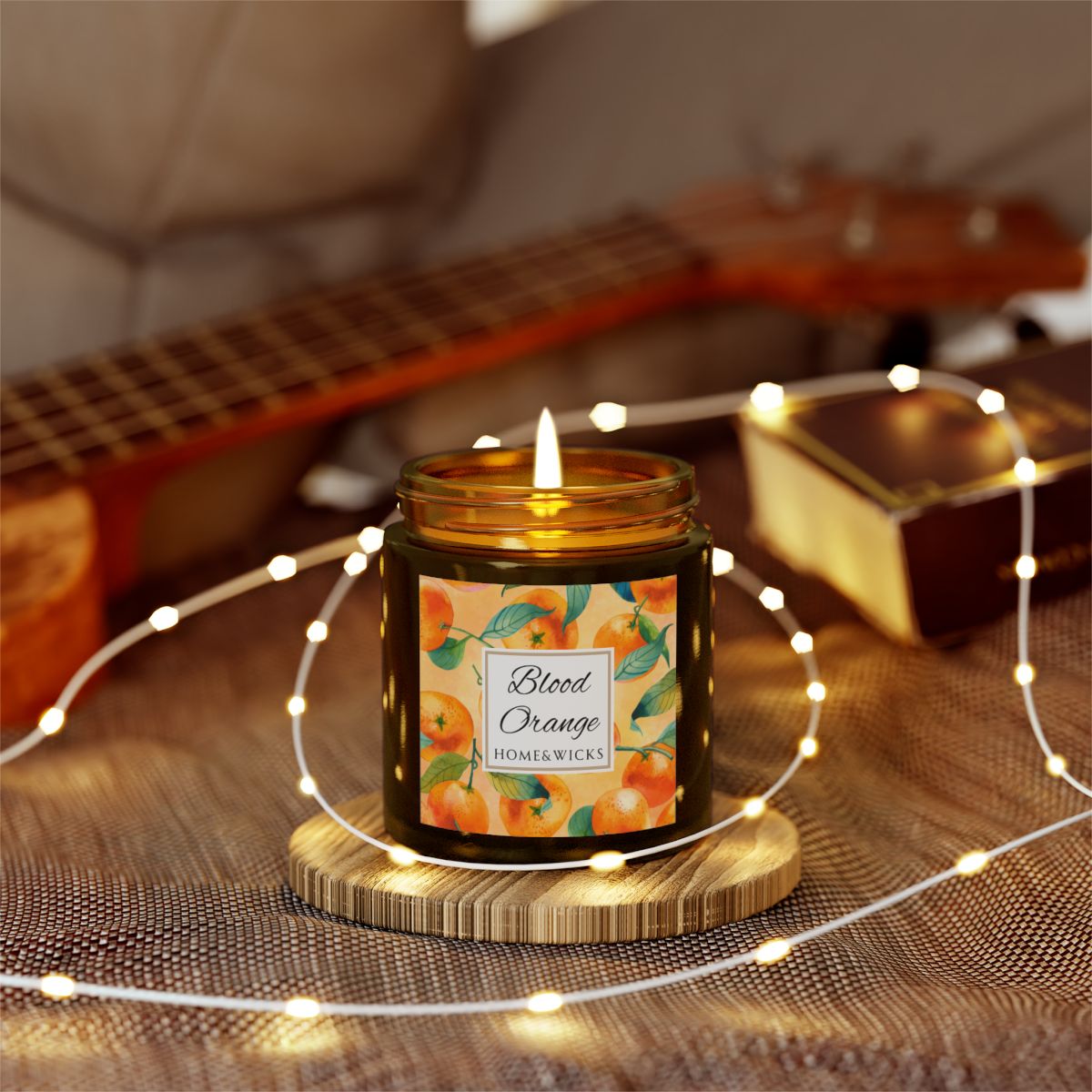 Blood Orange candle in an amber glass jar with gold lid with 'Blood Orange' label featuring oranges illustration on a white background by Home & Wicks on a wooden coaster surrounded by string lights, with a guitar in the background.