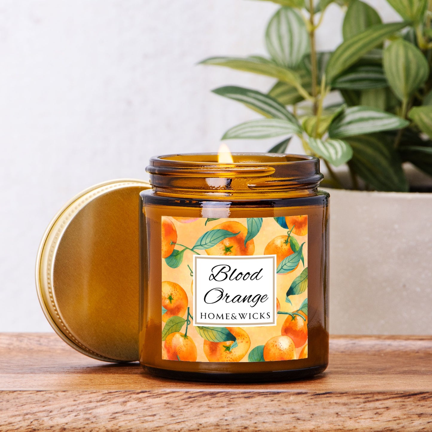 Blood Orange candle in an amber glass jar with gold lid with 'Blood Orange' label featuring oranges illustration on a white background by Home & Wickson a wooden surface with a plant in the background
