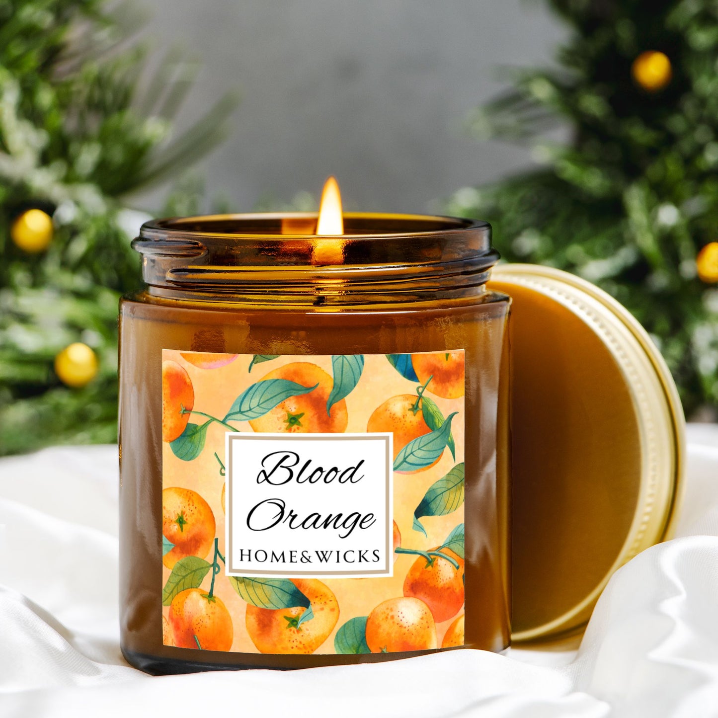 Blood Orange candle in an amber glass jar with gold lid with 'Blood Orange' label featuring oranges illustration on a white background by Home & Wicks surrounded by orange trees.