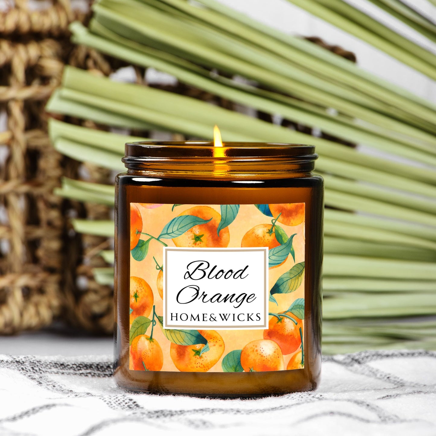 Blood Orange cotton wicked apricot coconut wax candle in an amber jar with gold lid and with 'Blood Orange' label and orange design on a white background by Home & Wicks on a textured surface with a woven basket and green leaves in the background.