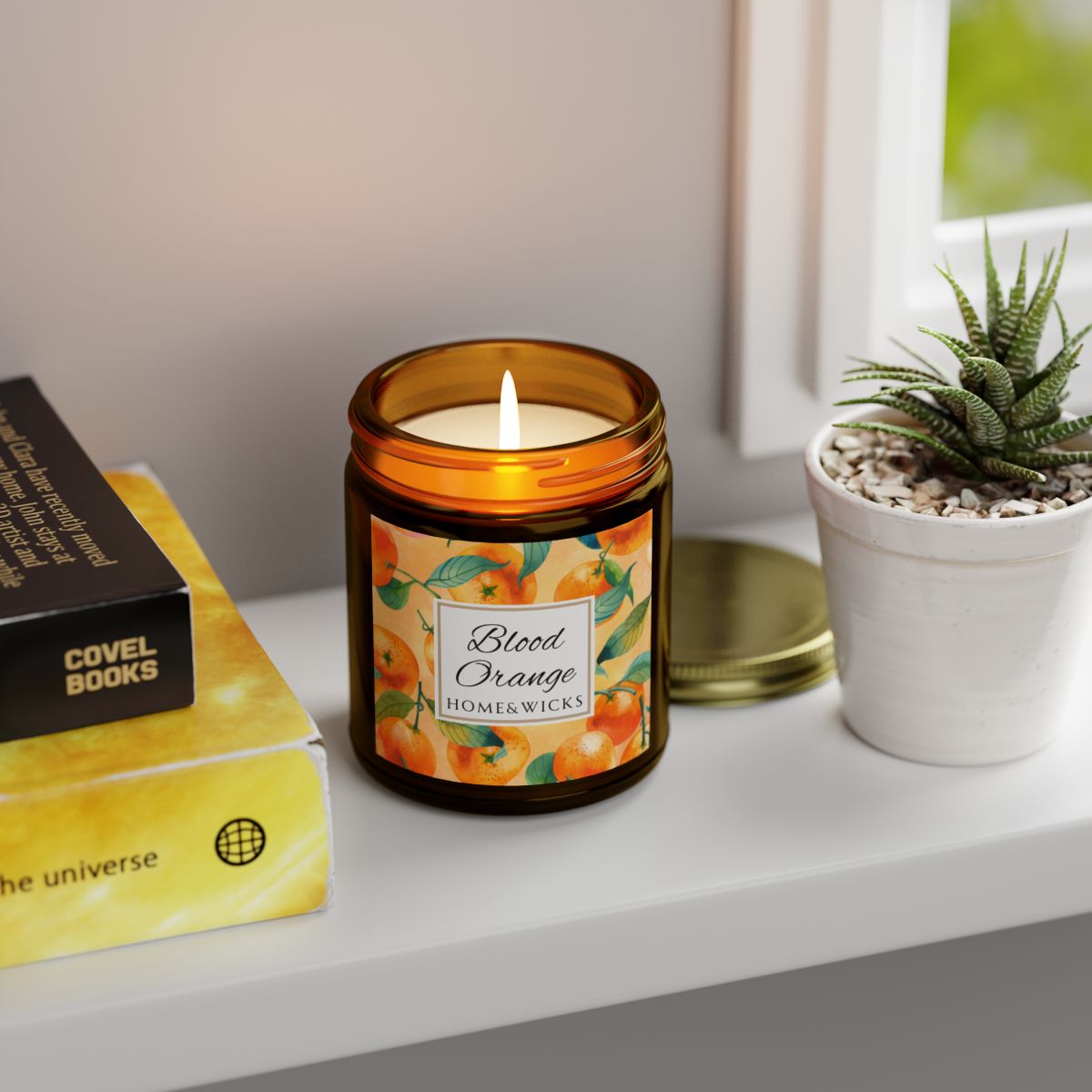 Blood Orange cotton wicked apricot coconut wax candle in an amber jar with gold lid and with 'Blood Orange' label and orange design on a white background by Home & Wicks on a shelf next to books and a plant.