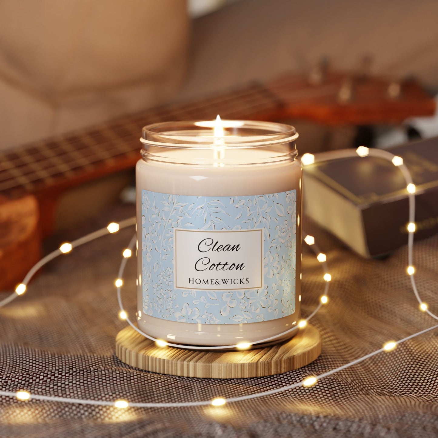 Candle labeled 'Clean Cotton' by Home & Wicks on a wooden coaster with string lights and a guitar in the background.