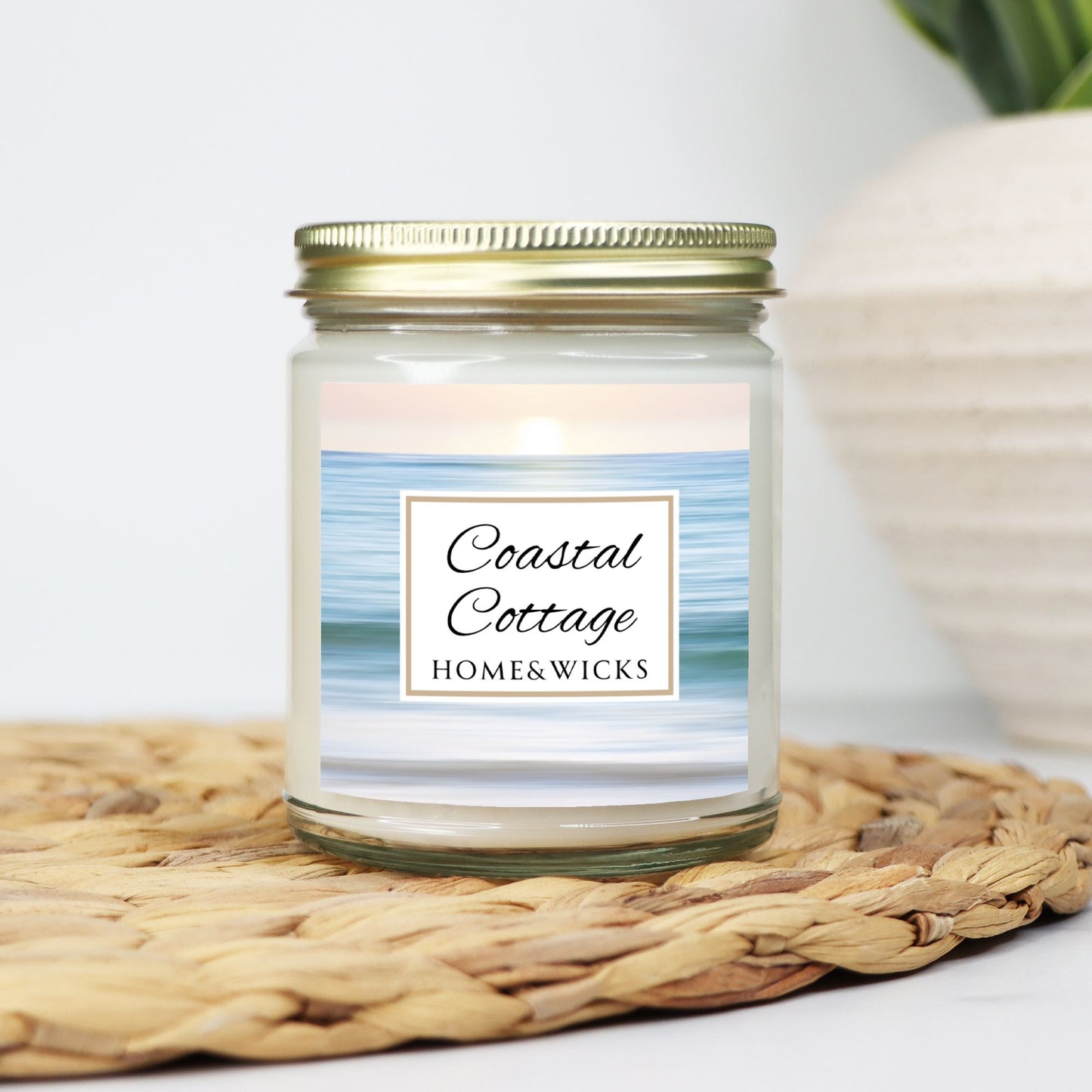 Coastal Cottage candle in clear jar with gold lid with a 'Coastal Cottage Home & Wicks' label with ocean shore image on a white background on a woven mat with a blurred background