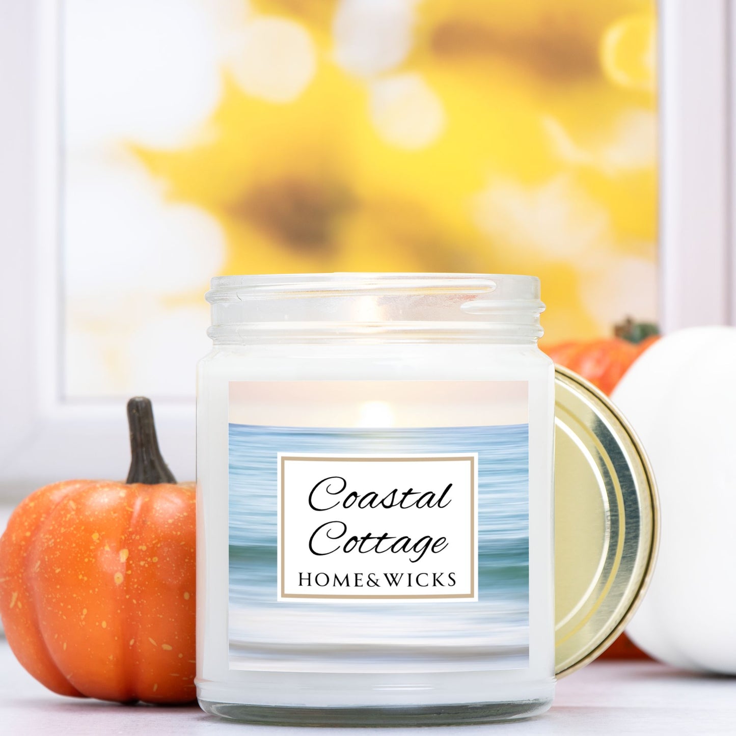 Coastal Cottage candle in clear jar with gold lid with a 'Coastal Cottage Home & Wicks' label with ocean shore image on a white background with pumpkins in the background