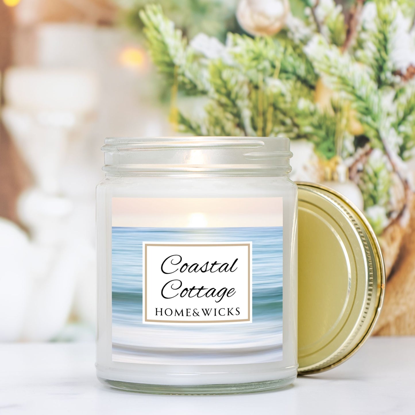 Coastal Cottage candle in clear jar with gold lid with a 'Coastal Cottage Home & Wicks' label with ocean shore image on a white background with greenery in background