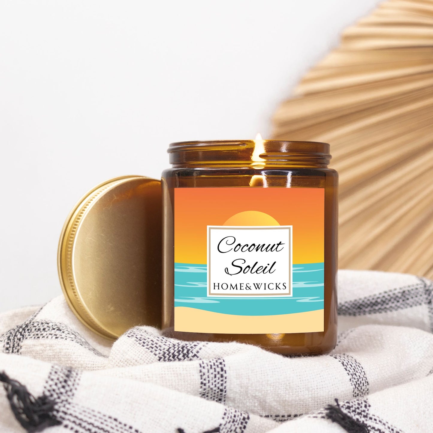Coconut Soleil coconut apricot wax candle in an amber jar with gold lid with a colorful ocean sunset graphic label featuring 'Coconut Soleil' by Home & Wicks on a white background.on a white and black patterned fabric with a wooden surface in the background.