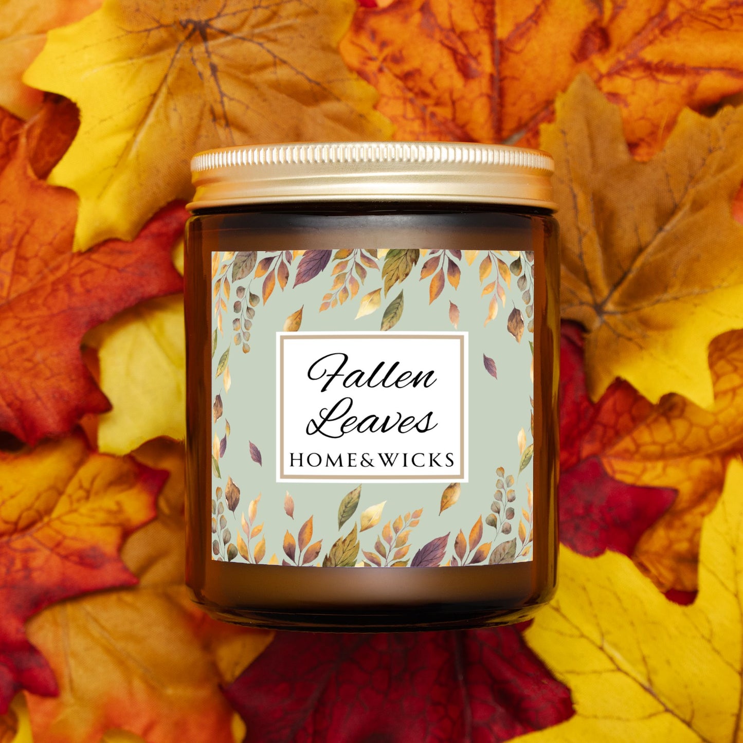 Amber glass candle jar with gold lid with a brown, gold and green 'Fallen Leaves' label on a white background by Home & Wicks on a bed of autumn leaves.