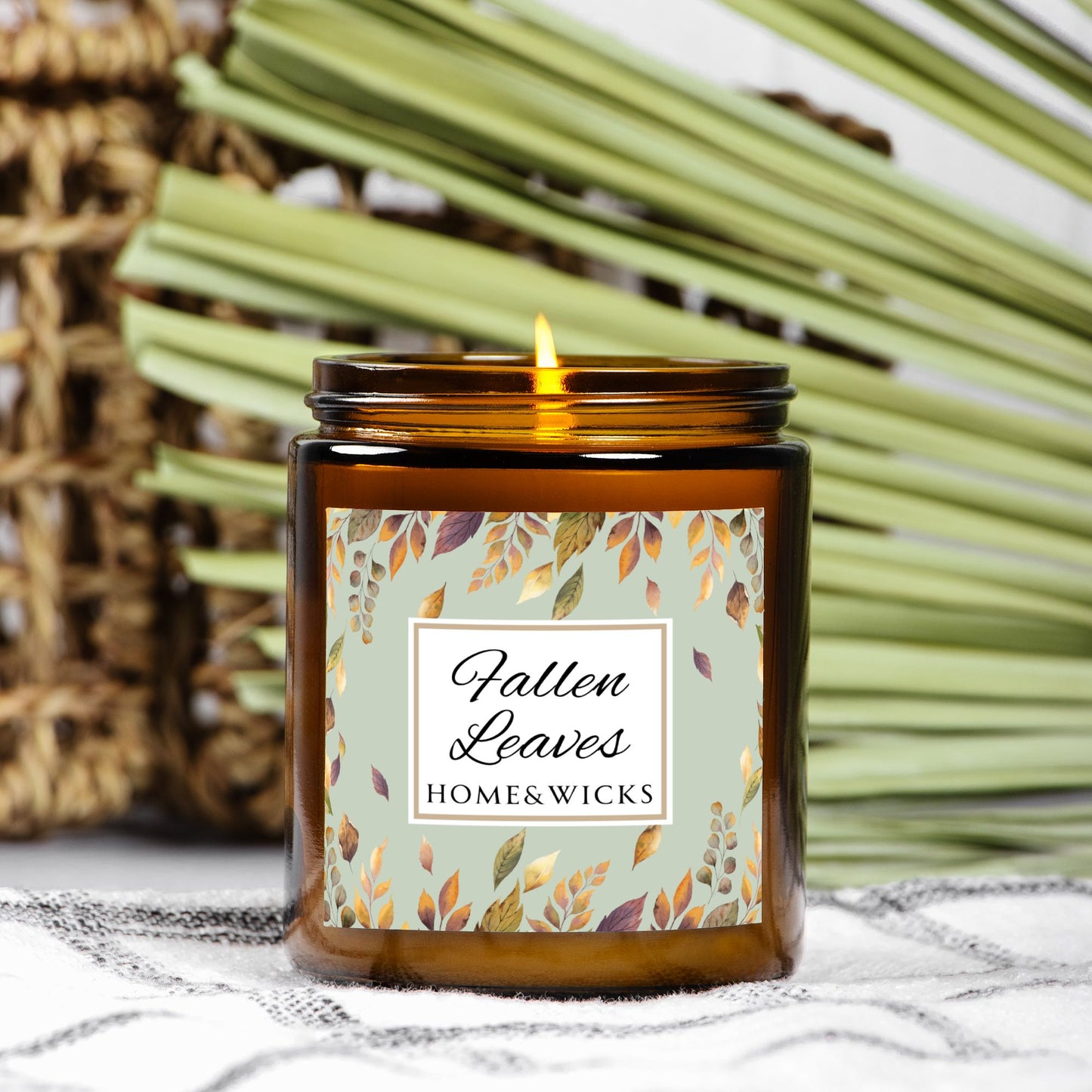 Amber glass candle jar with gold lid with brown, gold and green 'Fallen Leaves' label on a white background by Home & Wicks on a blanket with palm fran in background