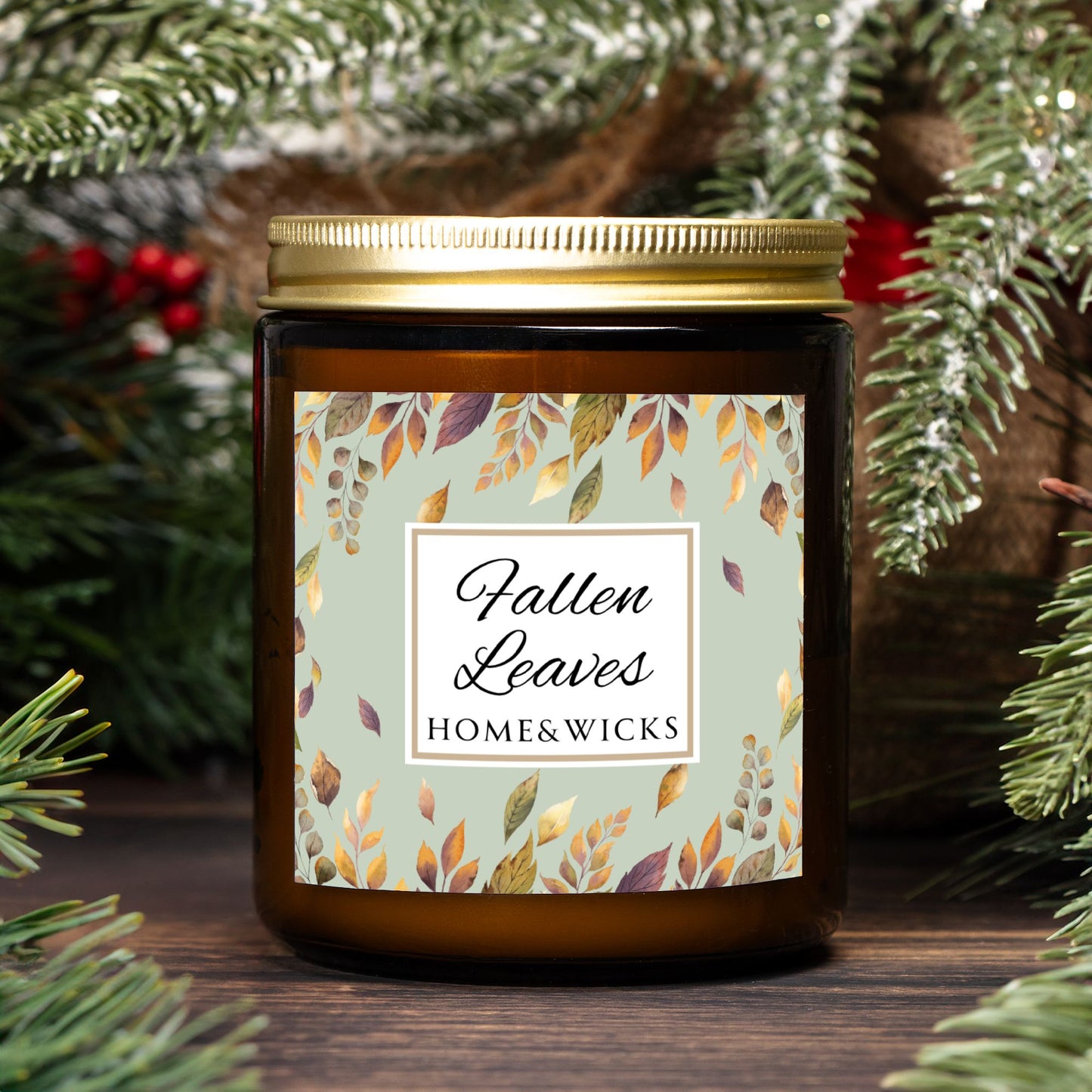 Amber glass candle jar with brown, gold and green 'Fallen Leaves' label on a white background by Home & Wicks, surrounded by greenery.