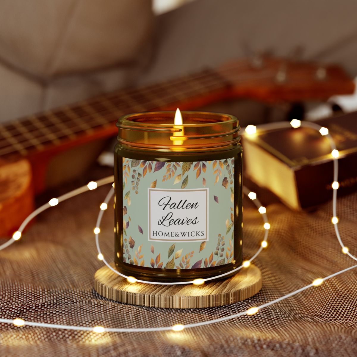 Amber glass candle jar with brown, gold and green 'Fallen Leaves' label on a white background by Home & Wicks, surrounded by string lights on a textured surface.