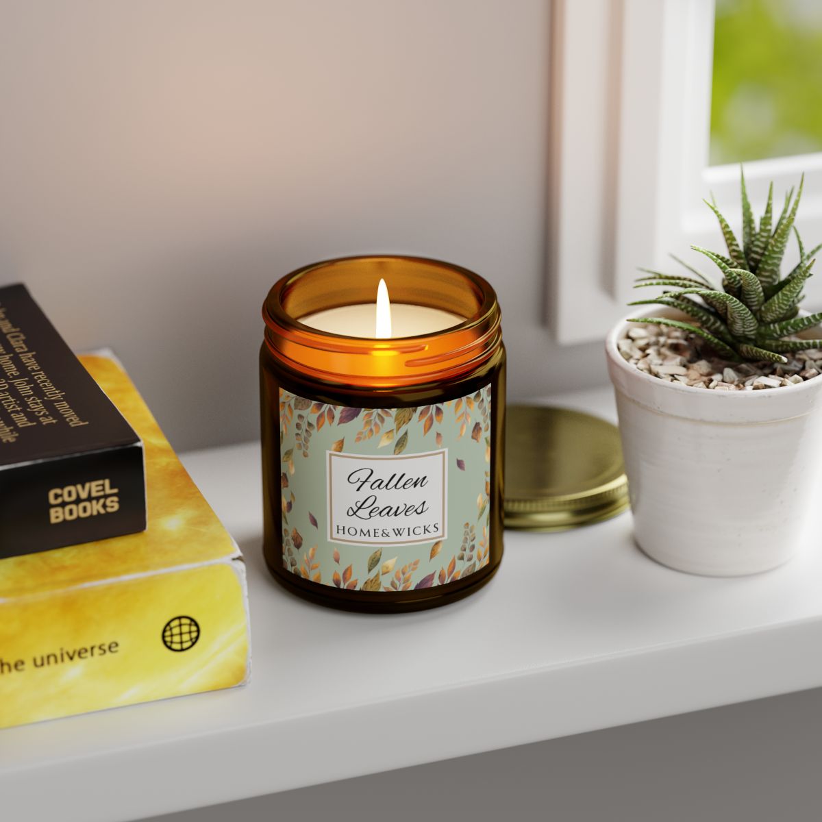 Amber glass candle jar with gold lid with brown, gold and green 'Fallen Leaves' label on a white background by Home & Wicks on a shelf with books and a plant.