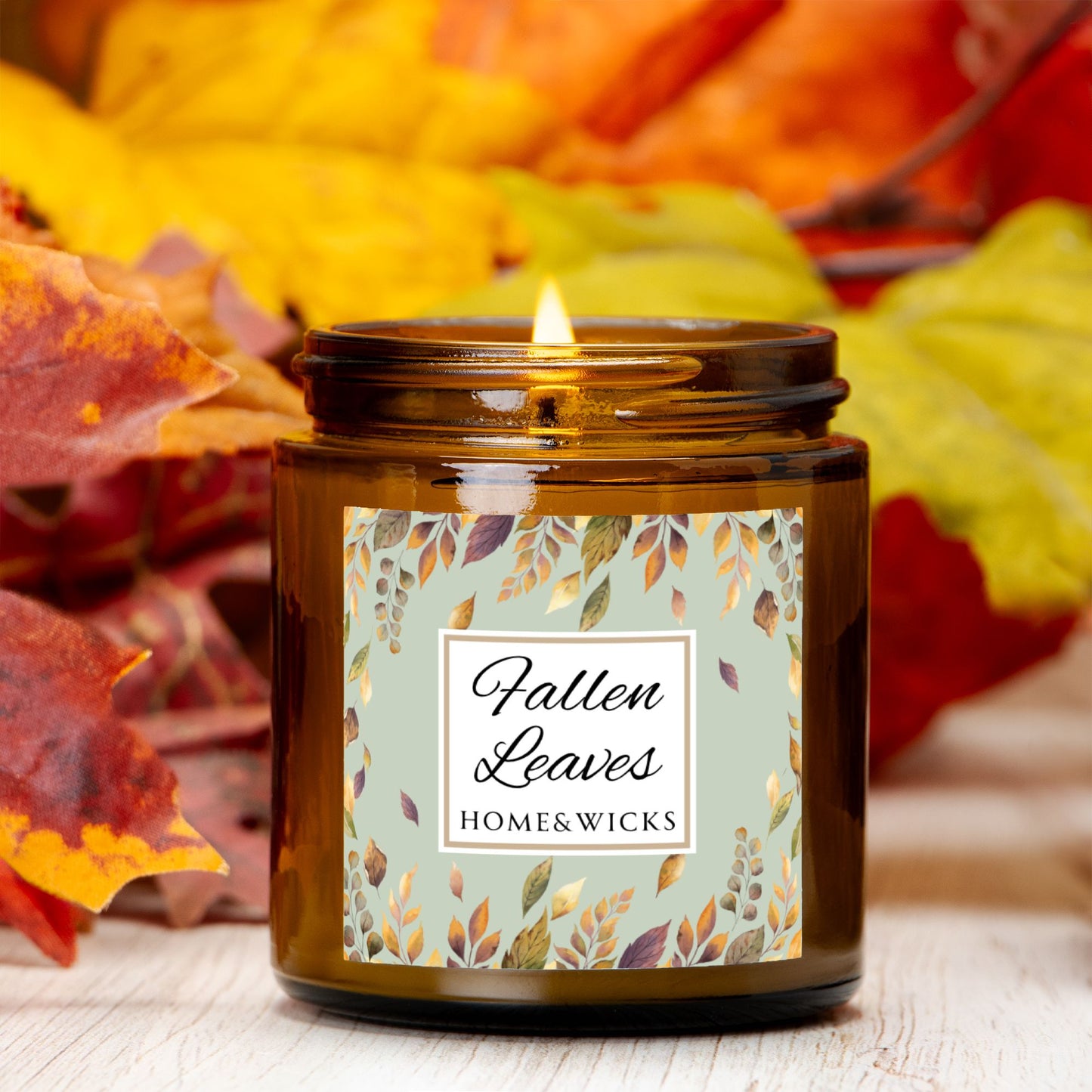 Fallen Leaves candle in a 4 oz. amber glass jar with gold lid with a 'Fallen Leaves' label on a white background by Home & Wicks with autumn leaves in the background.