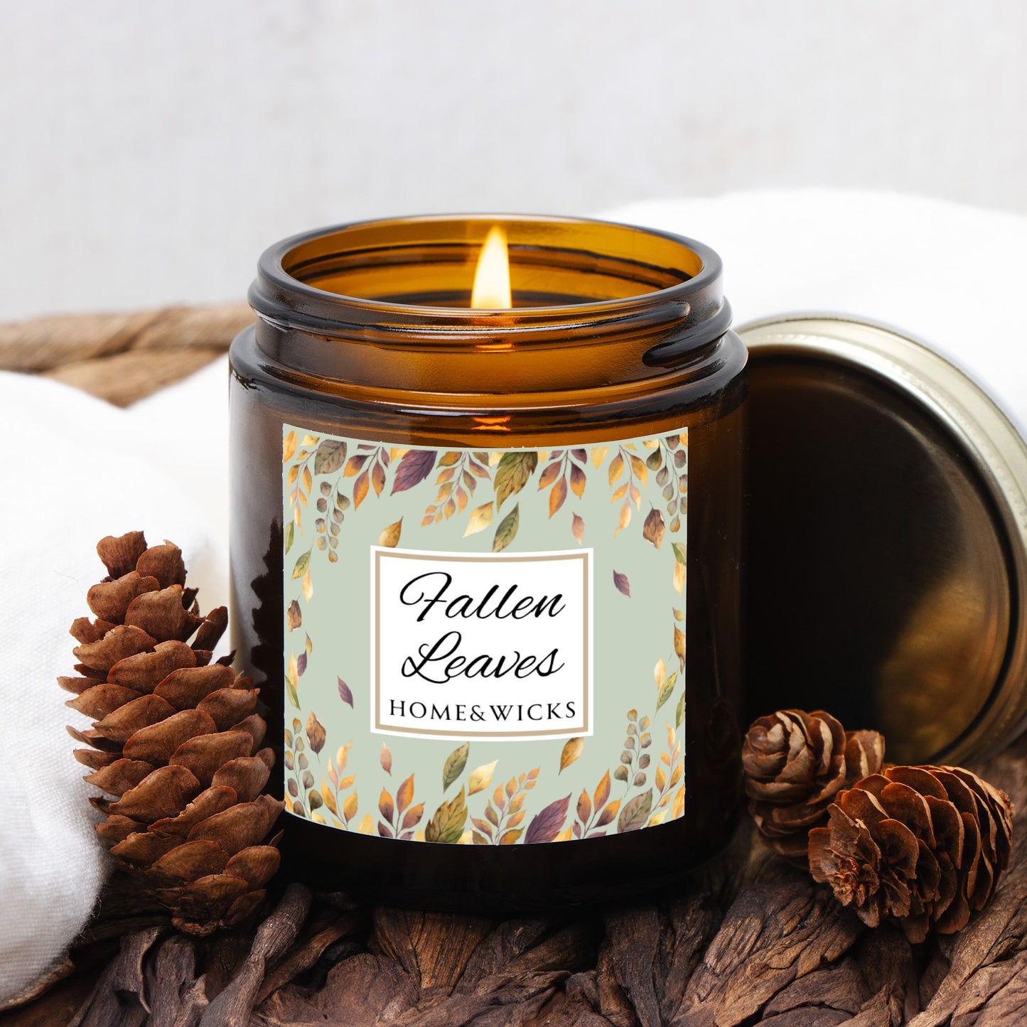 Fallen Leaves candle in a 4 oz. amber glass jar with gold lid with a 'Fallen Leaves' label on a white background by Home & Wicks surrounded by pine cones and wood on a white background.