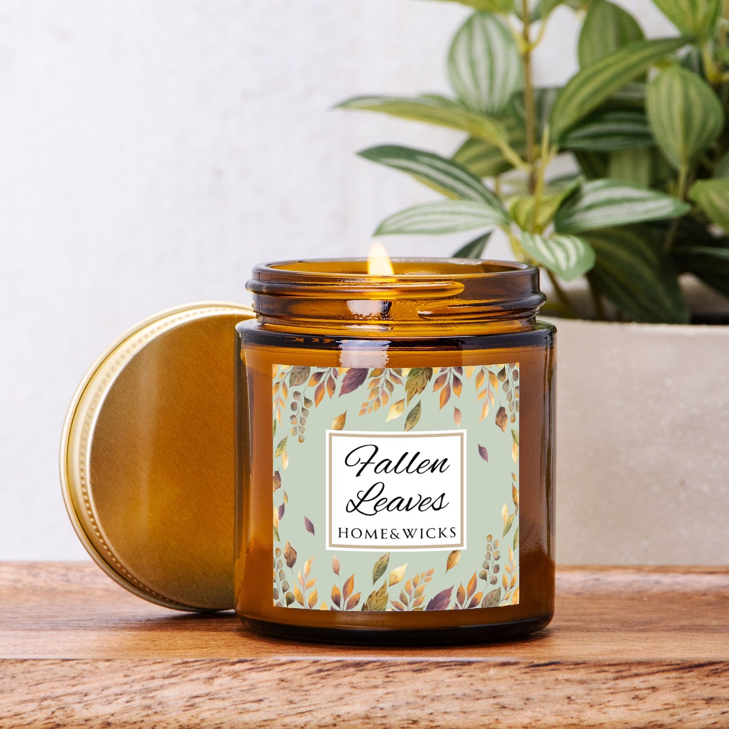 Fallen Leaves candle in a 4 oz. amber glass jar with gold lid with a 'Fallen Leaves' label on a white background by Home & Wicks on a wooden surface with a plant in the background.