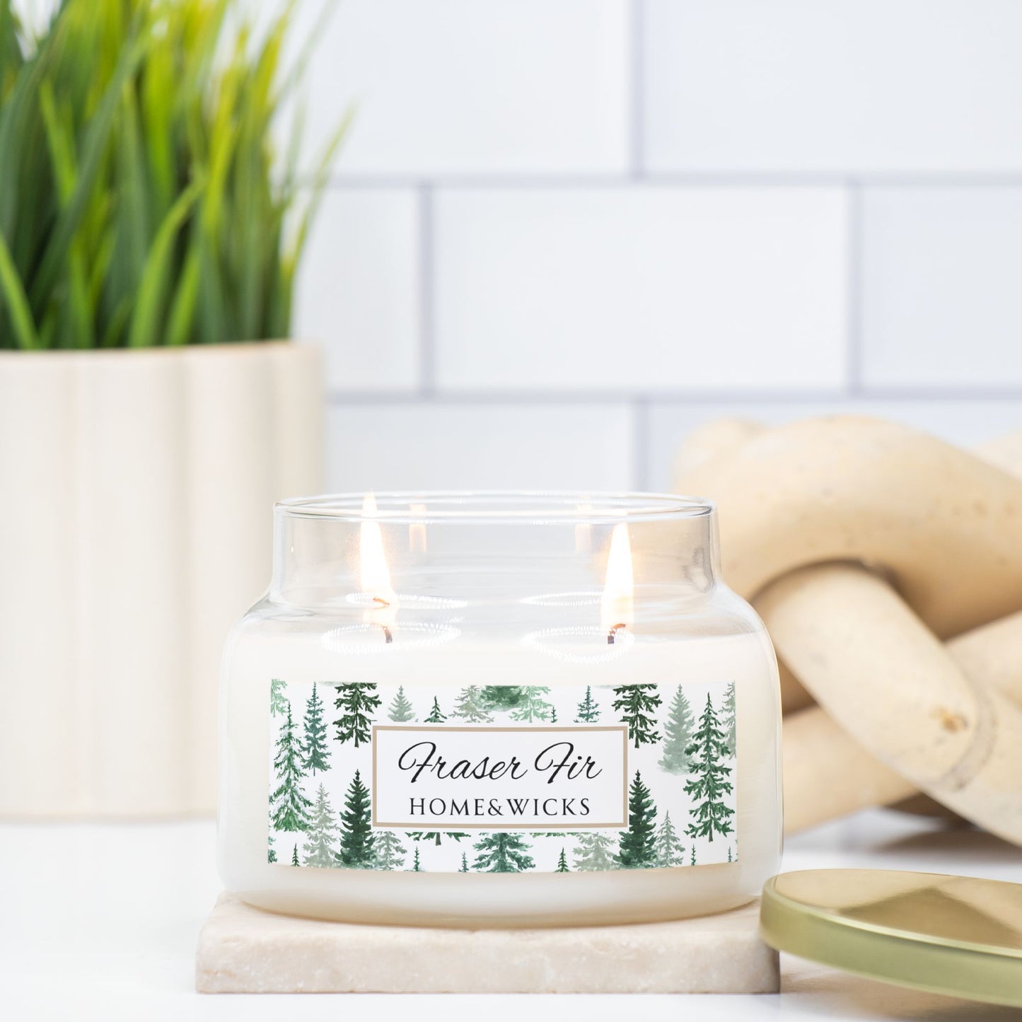 Candle with a 'Fraser Fir' label in a glass jar on a white surface with a plant and wooden logs in the background.