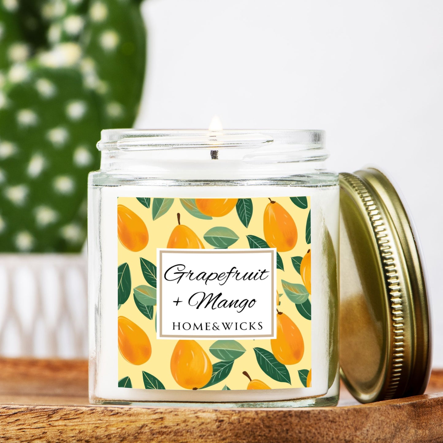 Grapefruit and Mango scented candle in a clear jar with gold lid with a decorative mangoes label on a white background by Home & Wicks on a wooden surface.