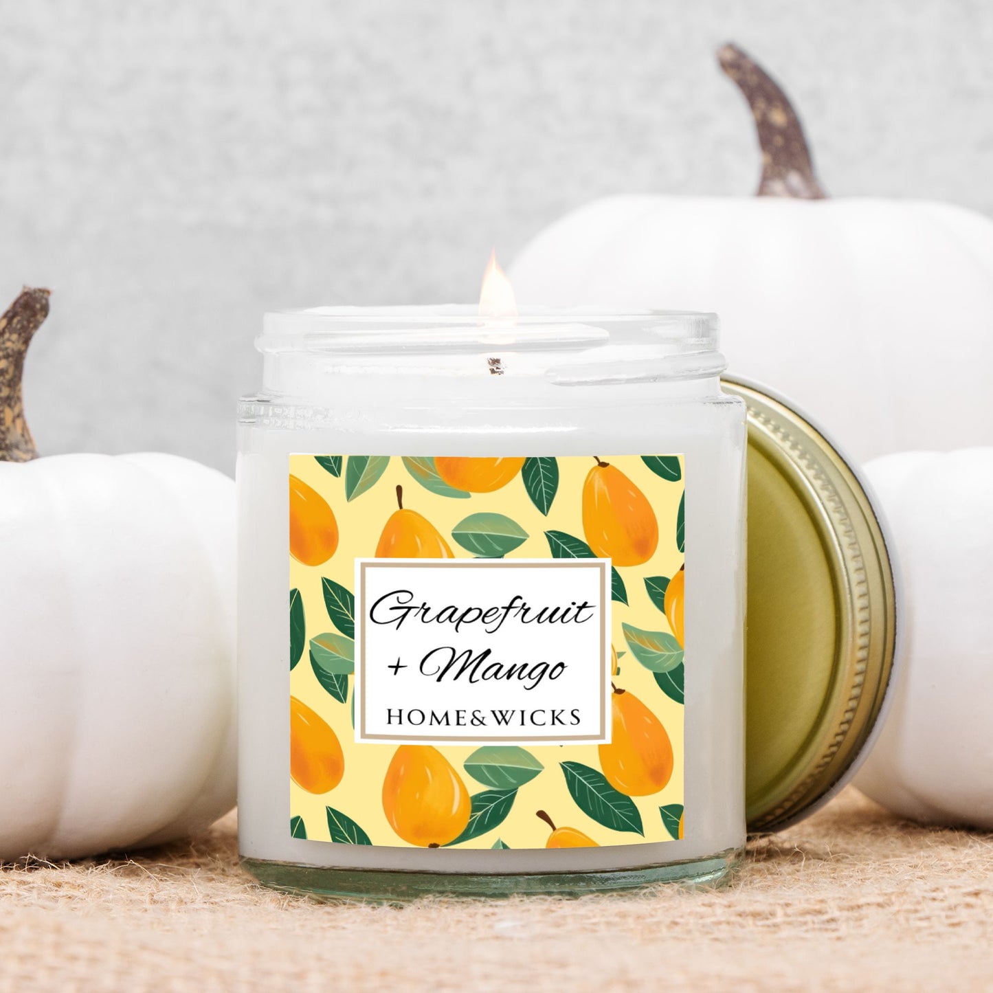 Grapefruit and Mango scented candle in a clear jar with gold lid with a decorative mangoes label on a white background by Home & Wicks in front of white pumpkins on a neutral background