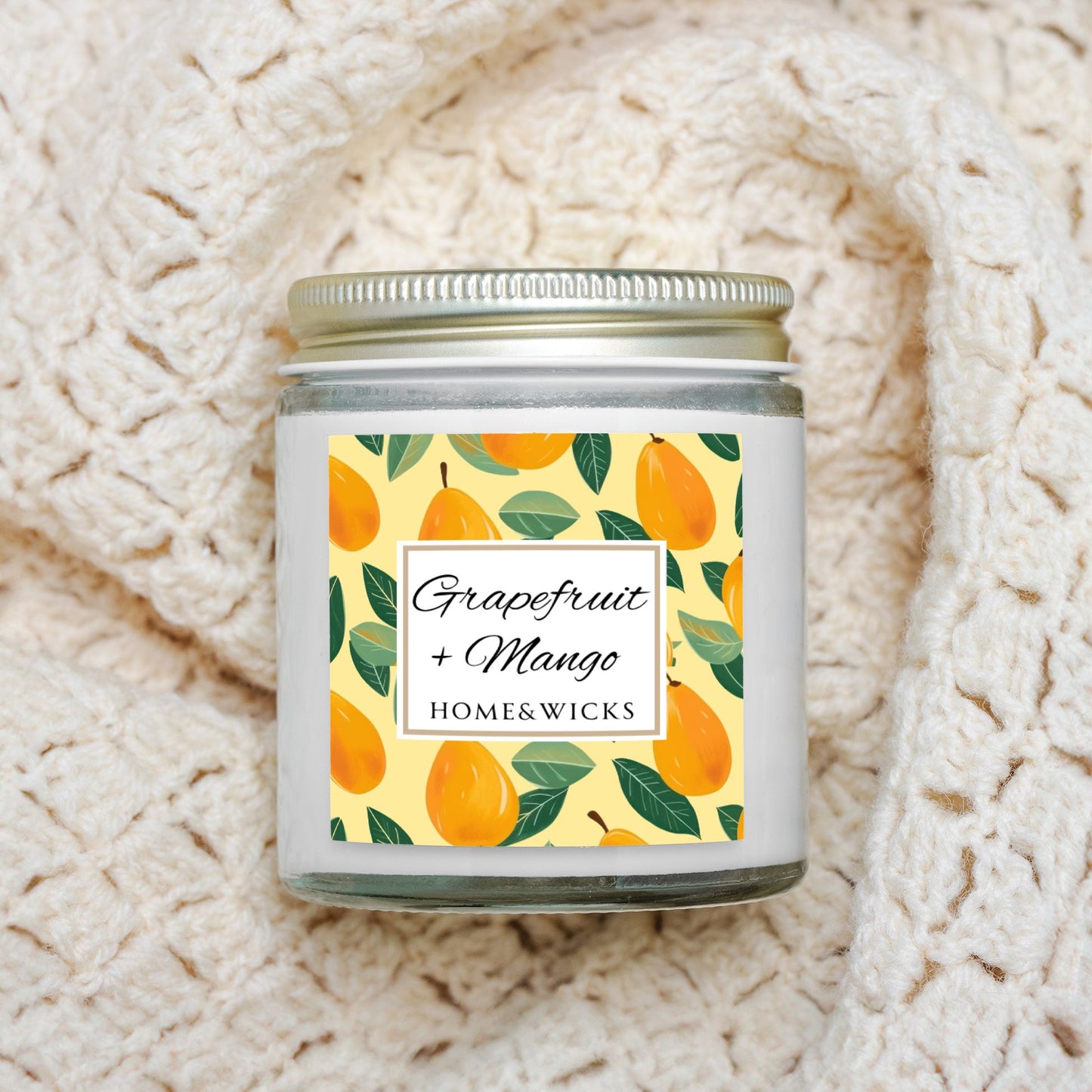 Grapefruit and Mango scented candle in a clear jar with gold lid with a decorative mangoes label by Home & Wicks on a textured beige blanket background