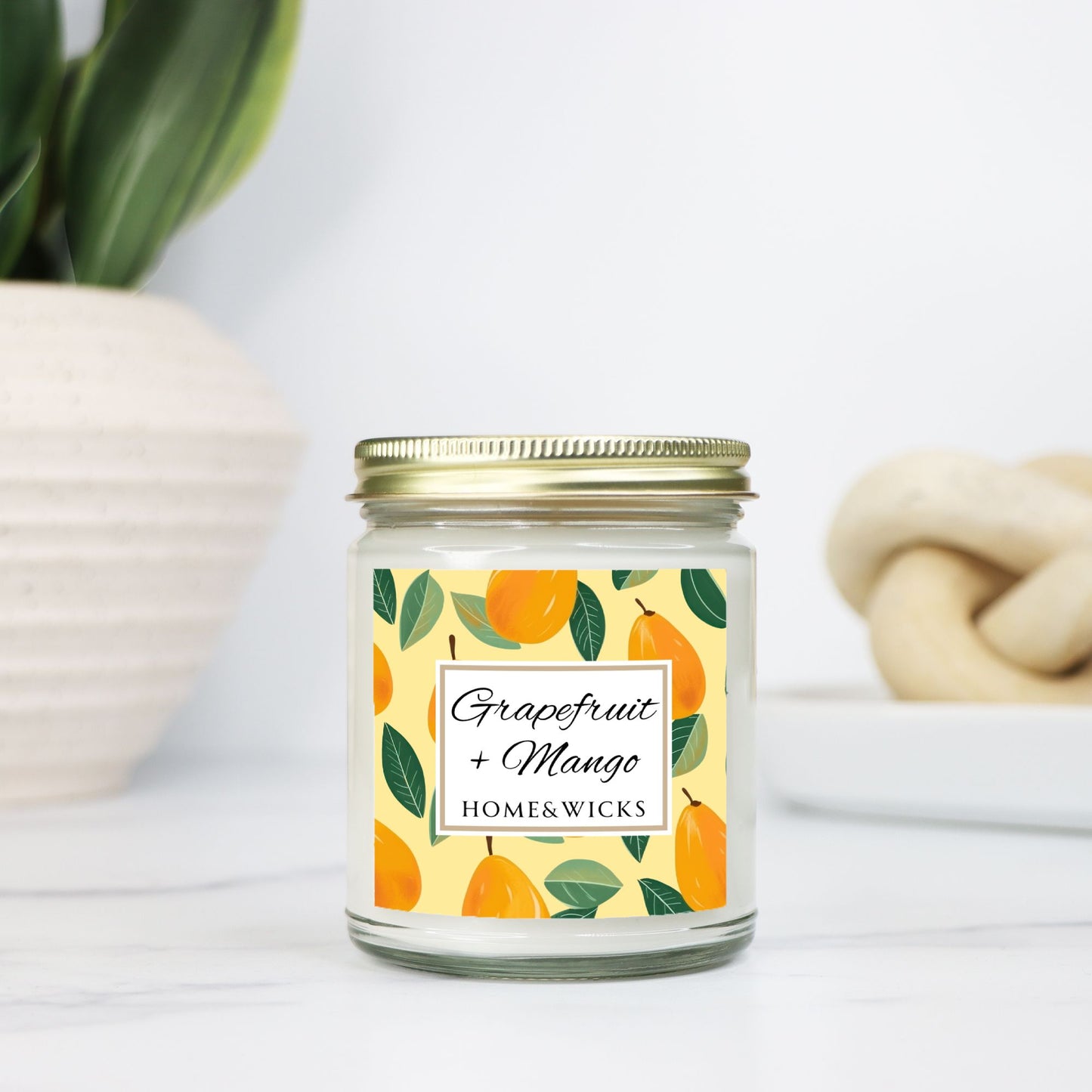 Grapefruit and Mango candle in a 9 oz. clear jar with gold lid and a decorative mango label on a white background by Home & Wicks on a light background