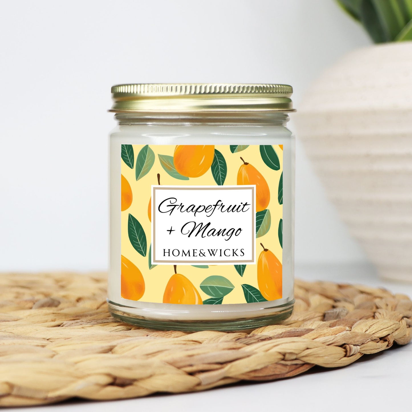 Grapefruit and Mango candle in a 9 oz. clear jar with gold lid and a decorative mango label on a white background by Home & Wicks on a woven mat.
