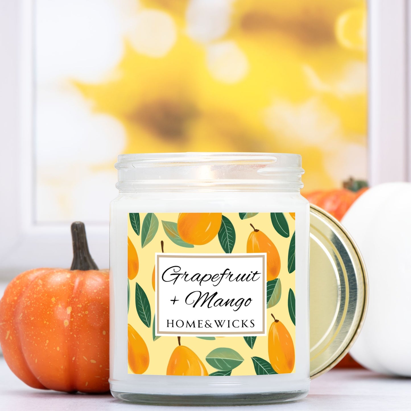 Grapefruit and Mango candle in a 9 oz. clear jar with gold lid and a decorative mango label on a white background by Home & Wicks on a blurred background with pumpkins