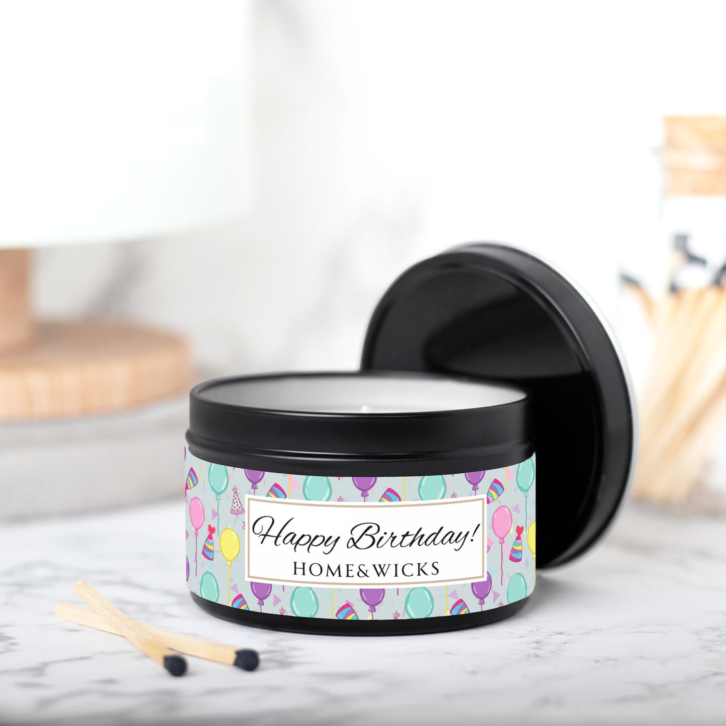 Black candle tin with 'Happy Birthday' label with colorful balloon, birthday hats and confetti design and 'Home & Wicks' branding on a light background.with match sticks