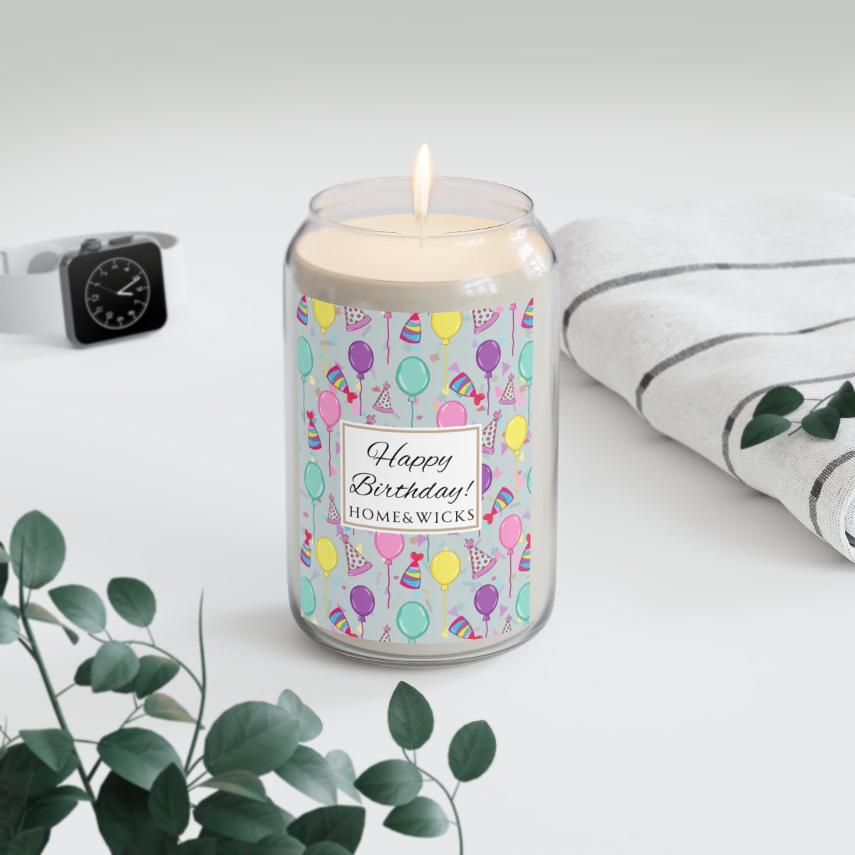 Large lit Happy Birthday 13.75 oz. Candle with birthday hats, confetti and balloons pattern with a blue-green light cyan background design on label on a white background with towel and eucalyptus branch