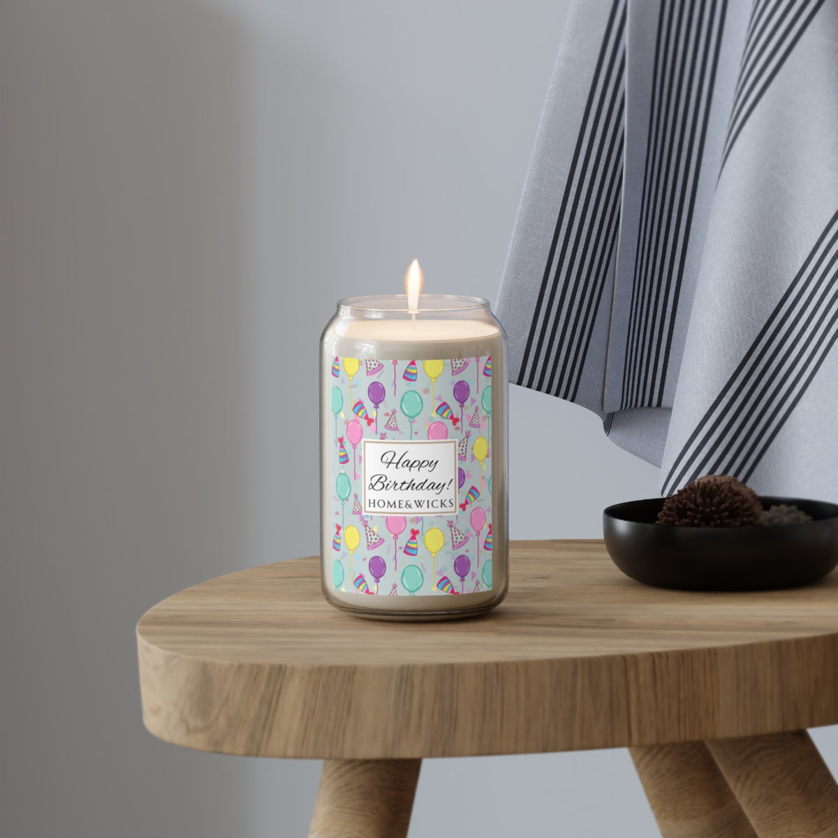 Large lit Happy Birthday 13.75 oz. Candle with birthday hats, confetti and balloons pattern with a blue-green light cyan background design on label on a white background sitting on wood table