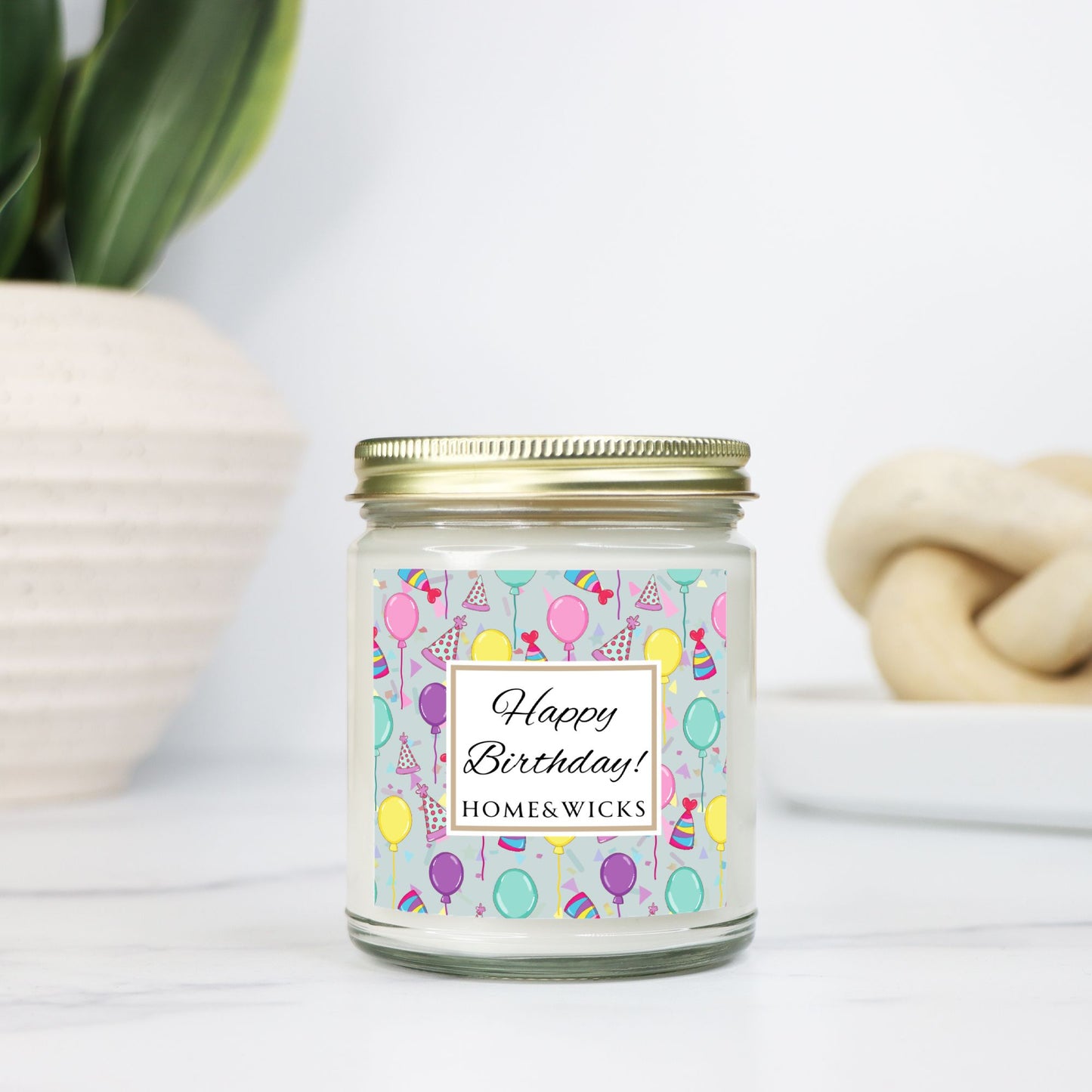 Candle in a clear jar with gold lid with a 'Happy Birthday!' with birthday hats, birthday balloons and confetti on label on a light background