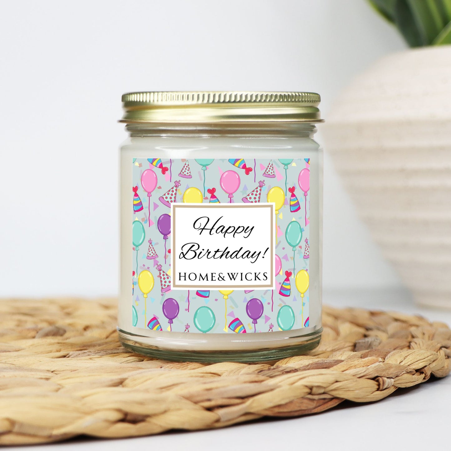 Candle in a clear jar with gold lid with a 'Happy Birthday!' with birthday hats, birthday balloons and confetti on label on a woven mat