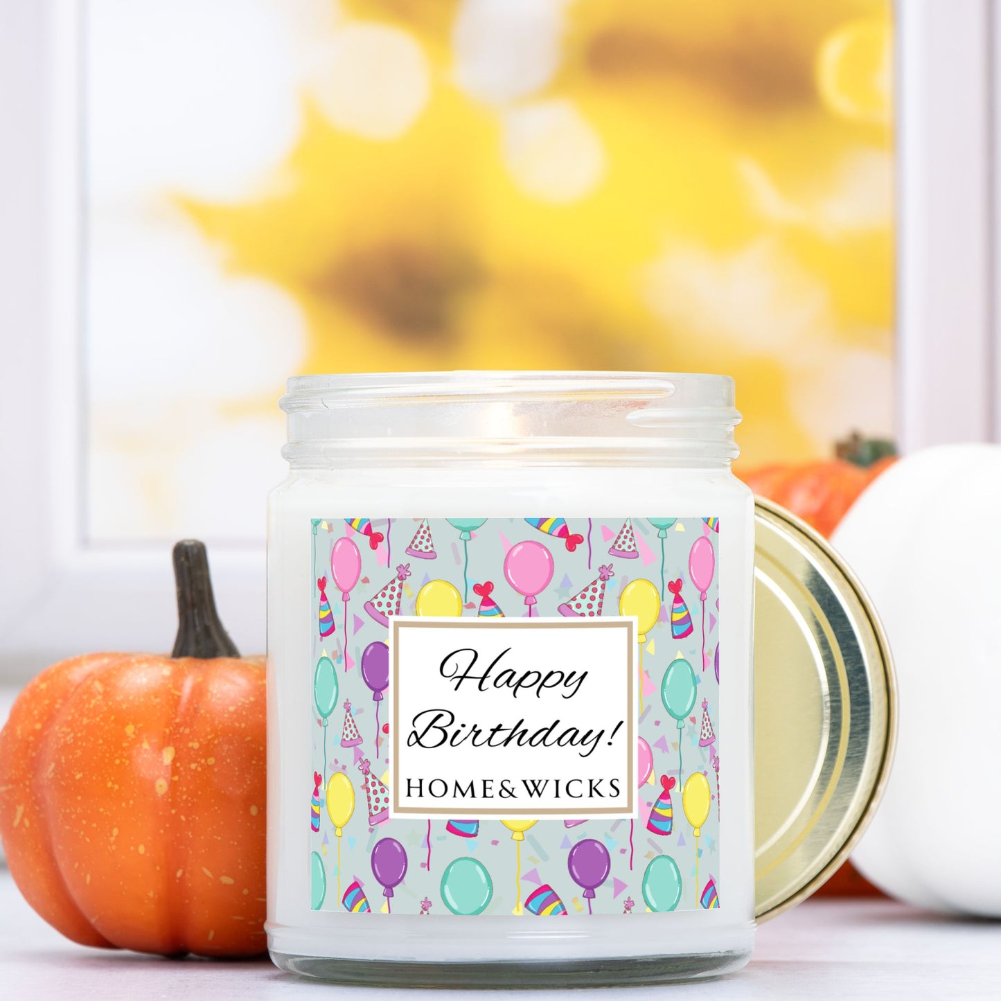 Candle in a clear jar with gold lid with a 'Happy Birthday!' with birthday hats, birthday balloons and confetti on label and pumpkins in the background