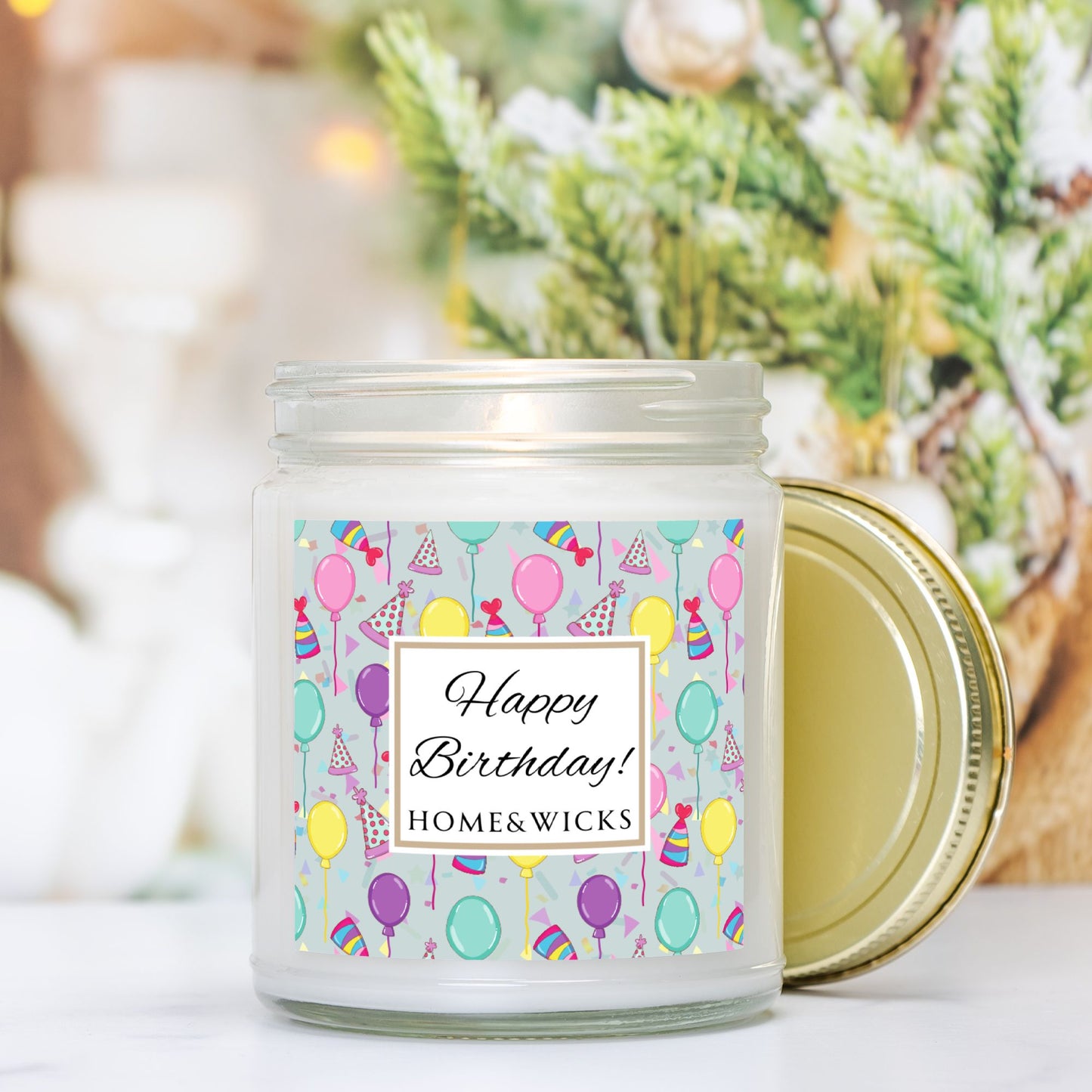 Candle in a clear jar with gold lid with a 'Happy Birthday!' with birthday hats, birthday balloons and confetti on label with greenery in background