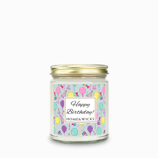 Candle in a clear jar with gold lid with a 'Happy Birthday!'  with birthday hats, birthday balloons and confetti on label on a white background