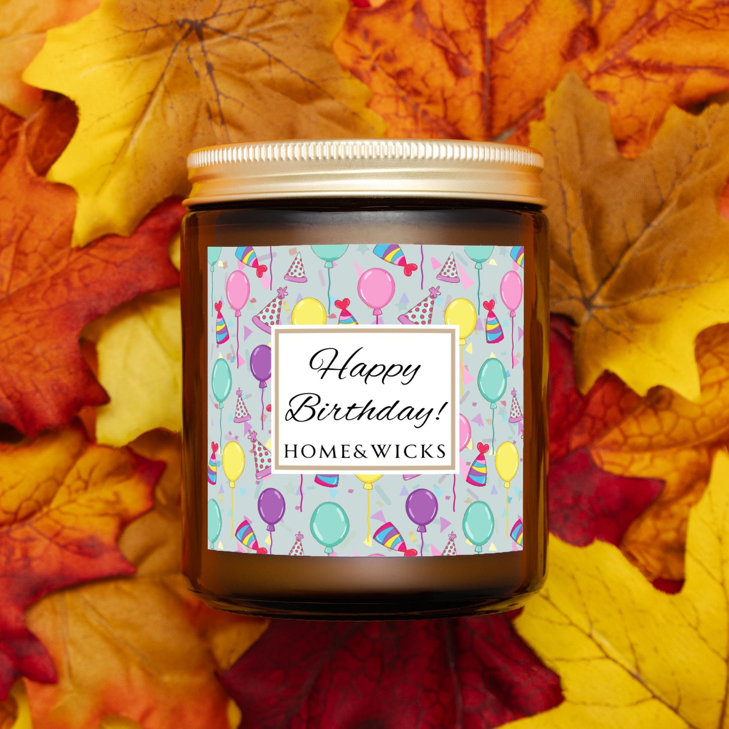 Amber candle jar with gold label with a 'Happy Birthday' label and colorful design with birthday balloons, birthday hats and confetti on a white background on a bed of autumn leaves