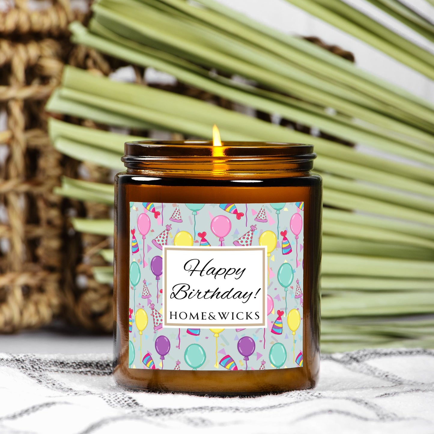 Amber candle jar with gold label with a 'Happy Birthday' label and colorful design with birthday balloons, birthday hats and confetti on a textured surface with wicker basket and green leaves in the background.