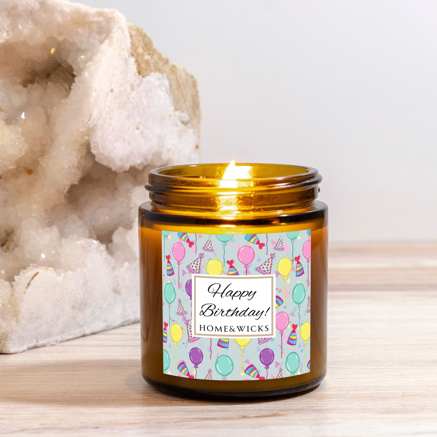 Amber candle jar with gold lid with a 'Happy Birthday' label featuring balloons, birthday party hats and confetti on a wooden surface with a white background